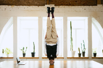 Office clerk having fun, doing headstand without hands on wooden table in modern office at work...
