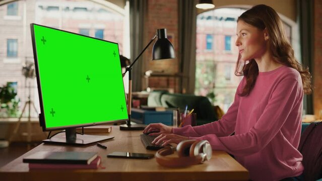 Young Beautiful Adult Woman Working from Home on Desktop Computer with Green Screen Mock Up Display. Creative Female Checking and Writing Emails. Loft Apartment with Urban City View from Big Window.