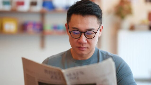 Concentrated Asian young man wearing eyeglasses reading newspaper in the cafe