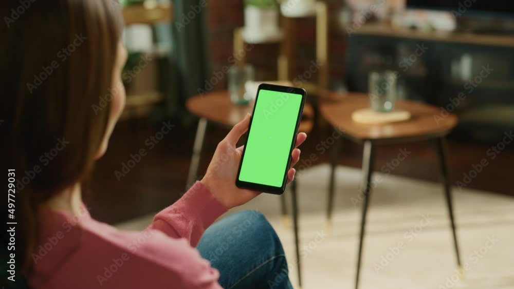 Poster feminine hand holding a smartphone with green screen mock up display. female is relaxing at home, wa