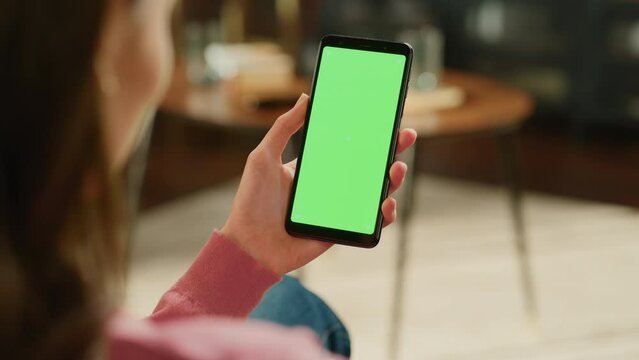 Feminine Hand Tapping on Display and Scrolling Feed on Smartphone with Green Screen Mock Up Display. Female Resting at Home, Checking Social Media on Mobile Device. Close Up Over the Shoulder Footage.