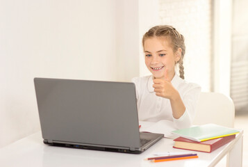 schoolgirl sitting at a table with a laptop and showing thumbs up