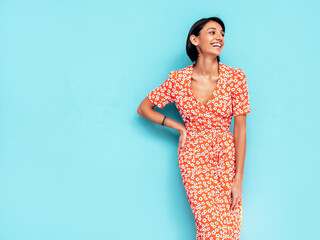 Obraz premium Young beautiful smiling female in trendy summer red dress. Sexy carefree woman posing near blue wall in studio. Positive model having fun. Cheerful and happy. Isolated