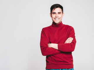 Portrait of handsome smiling model. Sexy stylish man dressed in red turtleneck sweater and jeans. Fashion hipster male posing on grey background in studio. Isolated. Crossed arms