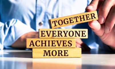 Team - together everyone achieves more sign on stacked wooden pegs wit businessman sitting in background.