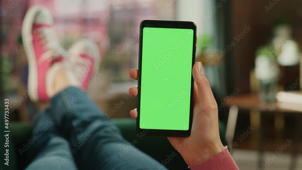 Aufkleber Feminine Hand Holding a Smartphone with Green Screen Mock Up Display. Female is Relaxing on a Couch at Home, Watching Videos and Reading Social Media Posts on Mobile Device. Close Up POV Footage.