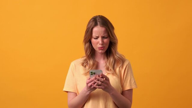 Displeased blonde woman wearing yellow t-shirt typing by phone in the orange studio