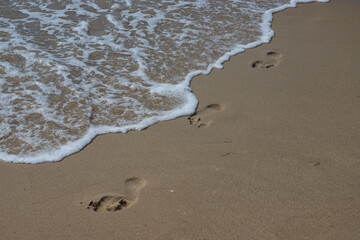 Footprints in the sand beach. Footprints in the sand against a sea wave. Footprints on a sunny day with golden sand, beach, wave and footsteps