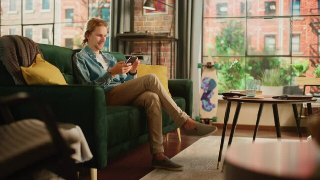 Young Handsome Adult Man Sitting on a Couch in Living Room, Relaxing and Using Smartphone. Creative Male Checking Social Media, Chatting with Friends, Browsing Internet. City View from Big Window.