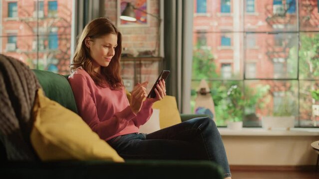 Young Beautiful Female Sitting on a Couch in Living Room, Relaxing and Using Smartphone. Creative Girl Checking Social Media, Chatting with Friends, Browsing Internet. City View from Big Window.