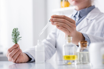 cropped view of laboratory assistant holding fir branch and pipette with serum near bottles in lab.