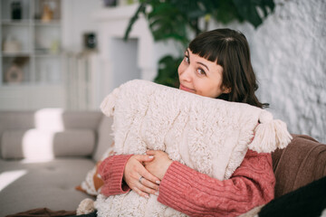 A young woman sits comfortably on a sofa and hugs a pillow in a modern bright apartment