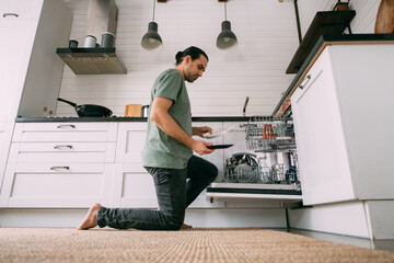 Weekend homework. A young man takes out clean dishes from the dishwasher in the kitchen at home.