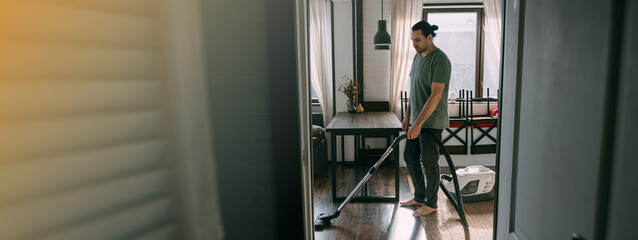 A young man is cleaning with a vacuum cleaner in the kitchen and living room at home.