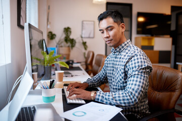 A businessman types on his computer while looking at printed report