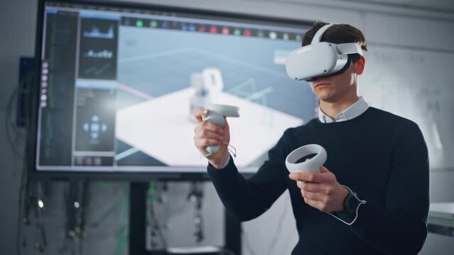 Futuristic University: Caucasian Male Engineer Wearing Virtual Reality Headset Uses Controllers to Remotely Moving Robotic Hand in Special App. Action of Robot Displayed on Screen