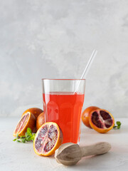 Red orange juice in a clear tall glass with a glass tube. Halves of orange on a light table. Mint leaves in the background. Light background