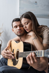 Fototapeta premium Young woman hugging boyfriend and playing acoustic guitar at home.
