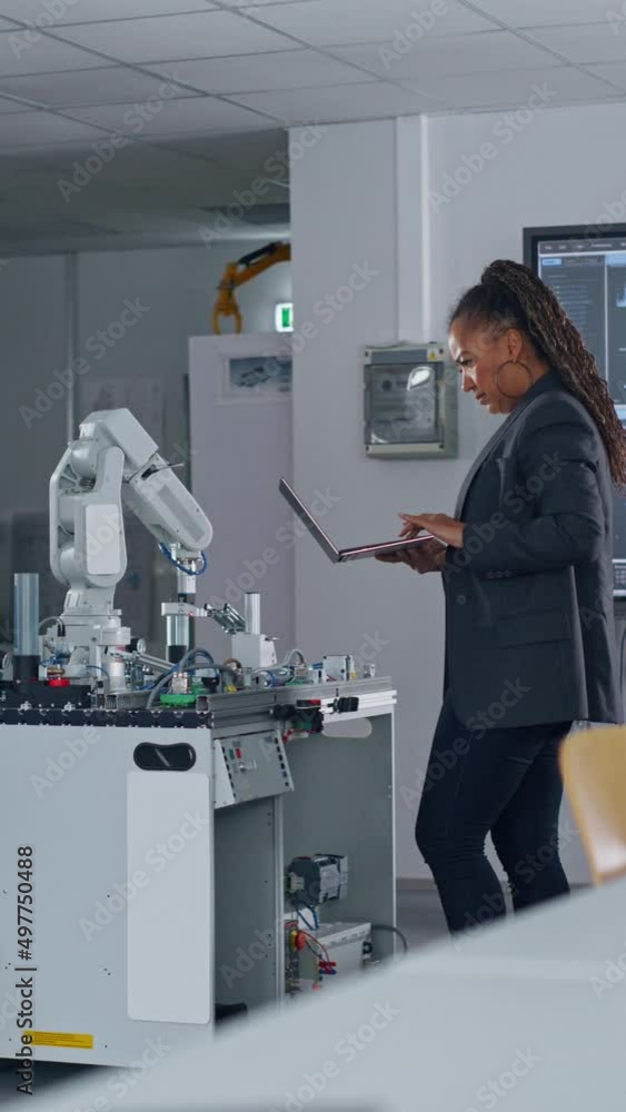 Poster At the Factory: Black Developer Uses Laptop for Programming Robotic Arm while Standing at Table In Front of Robot. New Era in Automatic Manufacturing Industry Concept. Vertical Screen