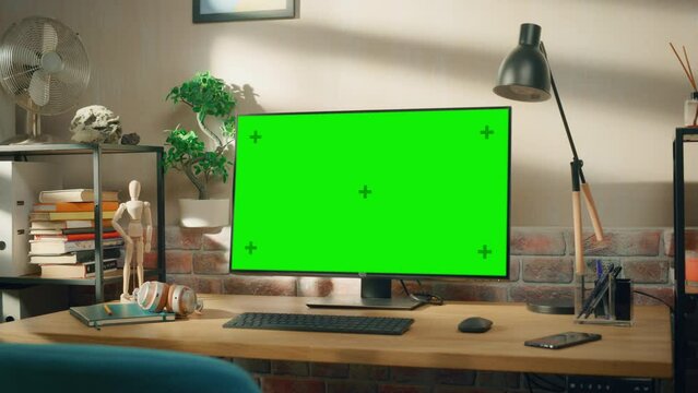 Desktop Computer Monitor Standing on a Desk with a Green Screen Chromakey Mock Up Display. Cozy Empty Loft Apartment with a Lamp, Notebooks and Smartphone on the Table. Zoom In Shot.