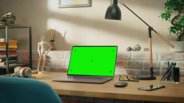 Laptop Computer Standing on a Wooden Desk with a Green Screen Chromakey Mock Up Display. Cozy Empty Loft Apartment with a Lamp, Notebooks and Smartphone on the Table. Zoom In Shot.