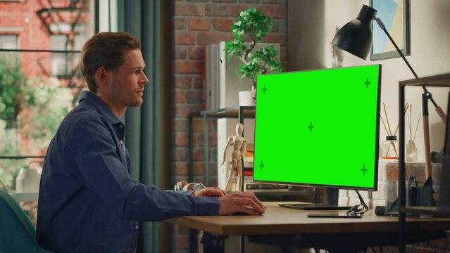 Young Handsome Man Working from Home on Desktop Computer with Green Screen Mock Up Display. Male Checking Corporate Accounts, Messaging Colleagues. Loft Living Room with Big Window. Arc Shot.