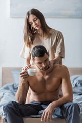 Fototapeta premium Blurred woman in t-shirt touching muscular man holding cup on bed.