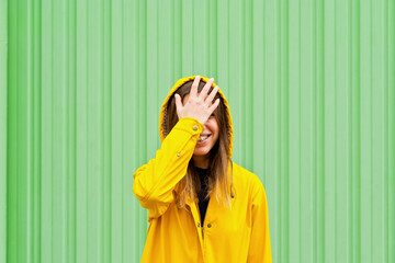 Mid waist view of woman covering her face in a yellow raincoat. Horizontal view of caucasian woman outdoors with yellow raincoat isolated on green wall. People isolated in background with copy space.