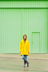 Vertical front view of woman wearing a yellow raincoat standing on the street. Full length body of woman under the rain with wellingtons and green facade. People isolated in background concept.