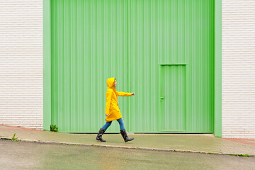 Horizontal side view of woman wearing a yellow raincoat walking on the street. Full length body of woman under the rain with wellingtons and green background. People isolated in background concept.