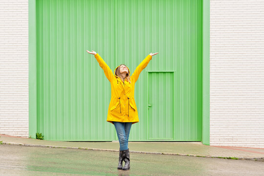 Horizontal front view of woman raising arms in a yellow raincoat standing on the street. Full length body of woman under the rain isolated on green wall. People isolated in background concept.