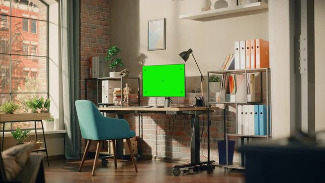 Desktop Computer Monitor Standing on Height Adjustable Desk with a Green Screen Chromakey Mock Up Display. Cozy Empty Loft Apartment with a Lamp, Notebooks and Smartphone on the Table. Wide Shot.