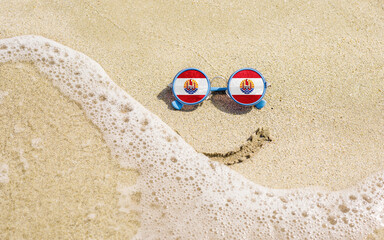 Sunglasses with flag of French Polynesia on a sandy beach. Nearby is a sea lightning and a painted smile. The concept of a successful vacation in the resorts of the French Polynesia.