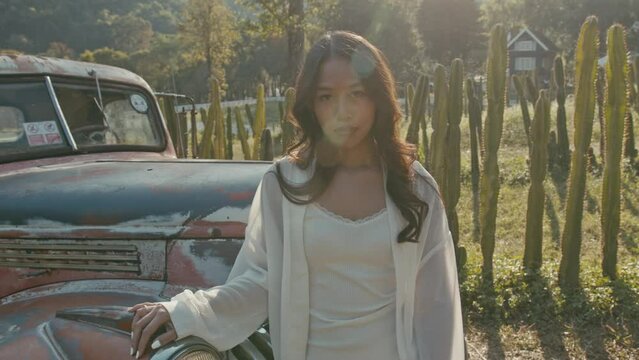 Young Asian woman stands by an old pickup truck at sunset and looks at the camera