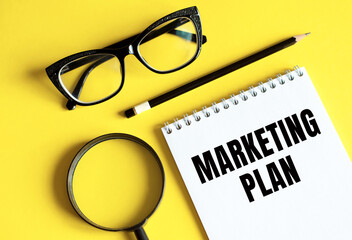 The phrase MARKETING PLAN in notebook with pencil, magnifier, and glasses on a yellow background