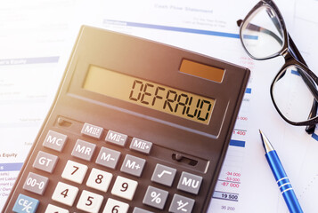 Defraud is a word written on calculator, lying on white surface next to glasses and a blue pen.