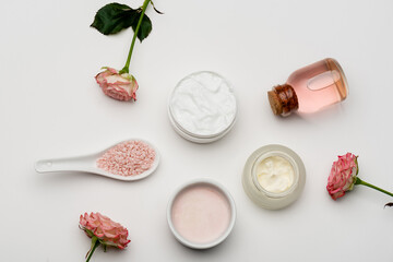 top view of flowers near pink sea salt, bottle with rose water and containers with cream on white.