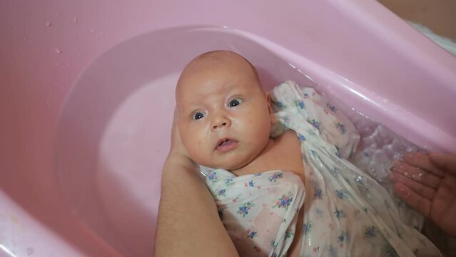 Mother splashes water on satisfied little baby washing in pink bath. Adorable newborn girl wrapped in swaddle diaper at home upper close view