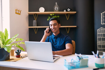 A wide shot of a young businessman taking a call at his desk in a trendy office