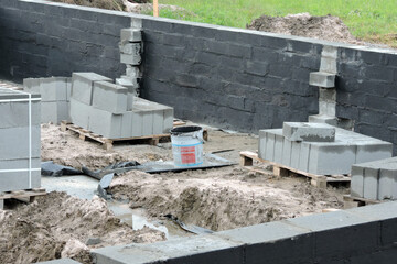 Damp proofing the outside foundation wall with tar, a construction site with solid concrete blocks on pallets, a plastic bucket