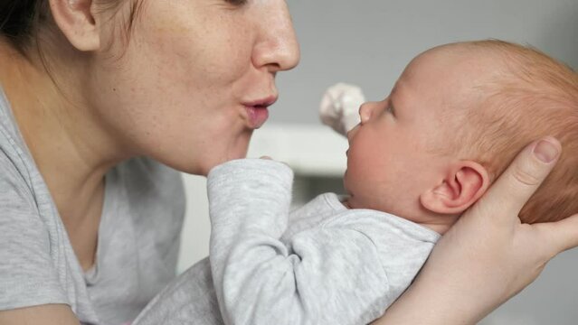 Happy mother babbles with newborn baby girl holding in arms on blurred background. Woman kisses face of cute little well-fed daughter at home closeup