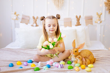easter, a little girl with painted colored eggs and a rabbit holding spring flowers tulips at home in a bright room preparing for the holiday smiling and having fun