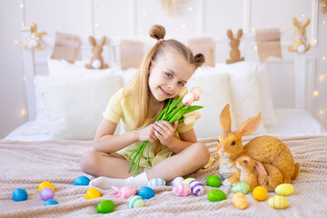 easter, a little girl with painted colored eggs and a rabbit holding spring flowers tulips at home in a bright room preparing for the holiday smiling and having fun