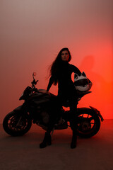Fototapeta na wymiar young teen brunette girl with long legs stands fashion with a biker helmet with ears near black sports motorcycle on a white wall background in red studio light. lifestyle fashion concept, free space