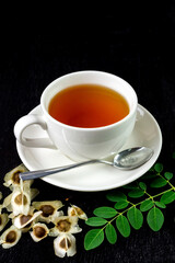 Moringa Tea in white cup with fresh green leaf and seed on black wooden background. Moringa oleifera tropical herb healthy lifestyle concept.