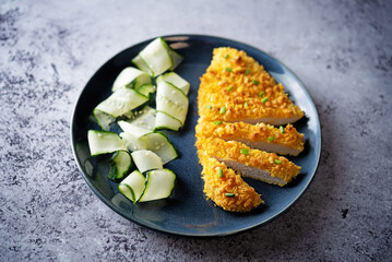 Bread crumbs cheesy chicken breast with cucumbers in a plate