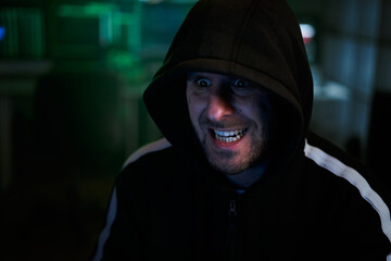Portrait of mysterious hooded hacker with evil face sitting at desk and breaking into government data servers in dark atmosphere with system codes animation in background.