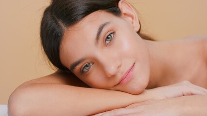 Close-up beauty portrait of young attractive brunette woman laying down her head on forearm and looks at camera against beige background