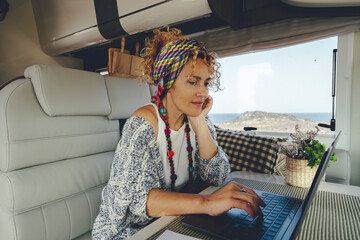Attracive young adult woman using laptop computer inside camper van alternative office. People and...