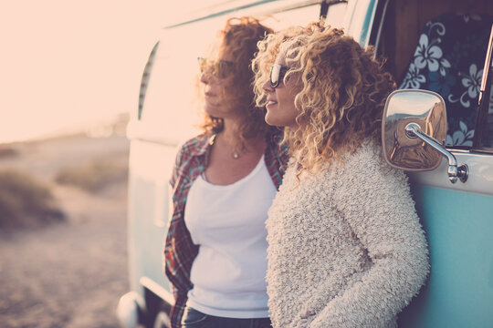 Two adult young woman enjoying time and leisure together against a classic blue van in travel vacation activity admiring the sunset. Friendship and female people traveler. Freedom emotion vanlife.